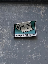 A sticker attached to a textured surface features a cartoonish skull with Xs for eyes and an open mouth, suggesting a humorous or deadpan expression. The background of the sticker is turquoise, and the word 'MALROLLITO' is printed at the bottom.