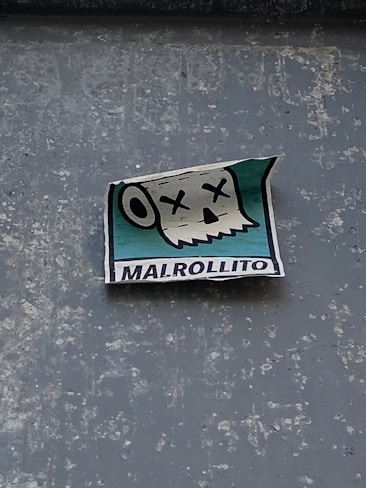 A sticker attached to a textured surface features a cartoonish skull with Xs for eyes and an open mouth, suggesting a humorous or deadpan expression. The background of the sticker is turquoise, and the word 'MALROLLITO' is printed at the bottom.