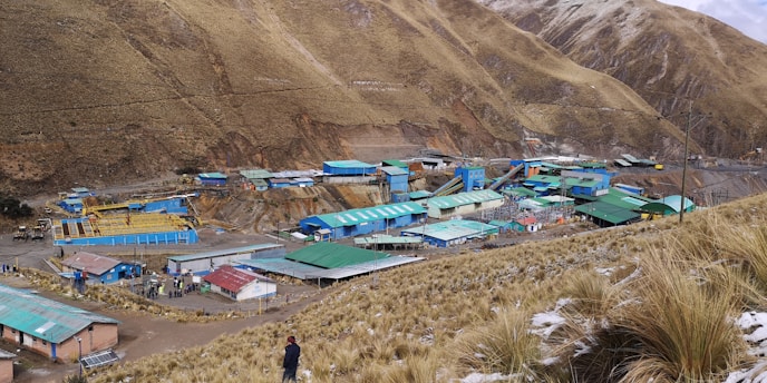 A rural area nestled in mountainous terrain with an industrial or mining setup. Various structures with green and blue rooftops are scattered across the landscape, surrounded by dry grass and rugged hills. A person stands in the foreground, looking towards the site.