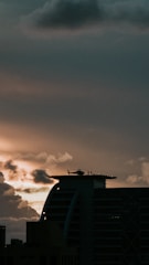 A helicopter is parked on top of a modern building with a helipad at sunset. The sky is filled with clouds and orange hues as the sun sets, casting a dramatic light over the scene. The silhouette of the building and helicopter contrasts against the colorful sky.