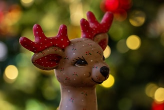A whimsical reindeer figurine with red antlers adorned with white polka dots, and small green and red decorative patterns on its face. The background consists of soft, out-of-focus circular light patterns in shades of green, yellow, and red, creating a festive atmosphere.
