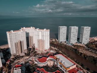 Aerial view of multiple high-rise apartment buildings situated near a waterfront, framed by a lush landscape with a river running in between. The ocean stretches out to the horizon under a partly cloudy sky, creating a tranquil atmosphere.