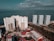 Aerial view of multiple high-rise apartment buildings situated near a waterfront, framed by a lush landscape with a river running in between. The ocean stretches out to the horizon under a partly cloudy sky, creating a tranquil atmosphere.
