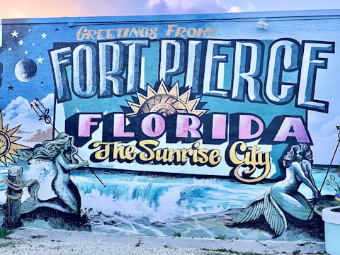 A colorful mural featuring the text 'Greetings from Fort Pierce Florida The Sunrise City' in large, bold letters. Surrounding the text are artistic depictions of mermaids, ocean waves, a sunburst, stars, clouds, and a crescent moon. The artwork includes intricate details and vibrant colors, capturing a coastal theme.