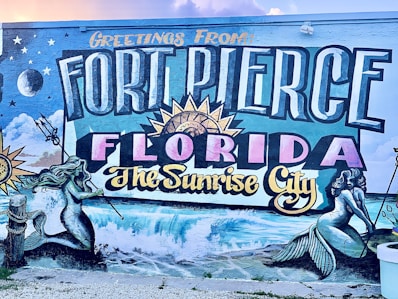 A colorful mural featuring the text 'Greetings from Fort Pierce Florida The Sunrise City' in large, bold letters. Surrounding the text are artistic depictions of mermaids, ocean waves, a sunburst, stars, clouds, and a crescent moon. The artwork includes intricate details and vibrant colors, capturing a coastal theme.