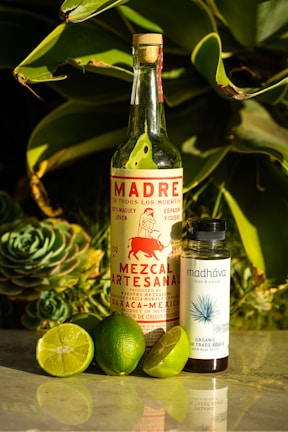 A bottle of artisanal mezcal labeled 'Madre de Todos Los Muertos' stands next to a smaller bottle of organic fair trade agave syrup. Surrounding them are several fresh limes, one of which is cut open. The background features lush green succulent leaves, creating a vibrant and natural setting.