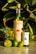 A bottle of artisanal mezcal labeled 'Madre de Todos Los Muertos' stands next to a smaller bottle of organic fair trade agave syrup. Surrounding them are several fresh limes, one of which is cut open. The background features lush green succulent leaves, creating a vibrant and natural setting.