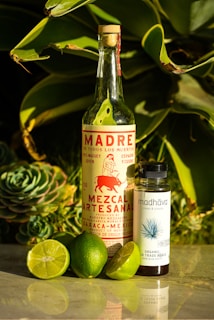 A bottle of artisanal mezcal labeled 'Madre de Todos Los Muertos' stands next to a smaller bottle of organic fair trade agave syrup. Surrounding them are several fresh limes, one of which is cut open. The background features lush green succulent leaves, creating a vibrant and natural setting.