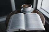 A quiet café table with a steaming cup of coffee and an open book by G Jamie.