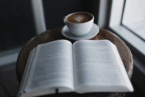 A quiet café table with a steaming cup of coffee and an open book by G Jamie.