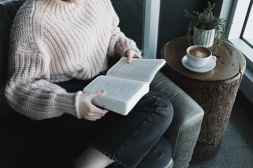 白いニットセーターとブルーデニムジーンズの女性が灰色のソファに座って本を読んでいます