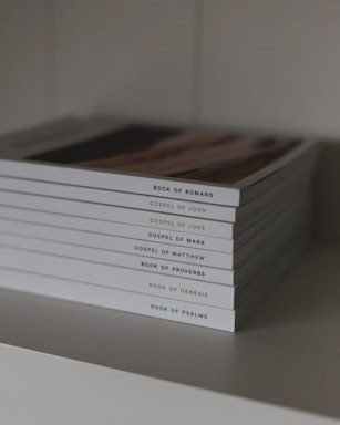 A stack of neatly arranged religious books with white covers displayed on a shelf, each titled with a different book of the Bible.