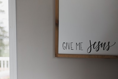 A bright, sunlit room features a white wall with a wooden-framed sign displaying the words 'Give Me Jesus' in a combination of capitalized and cursive fonts. On the left side, an out-of-focus window suggests an outdoor view.