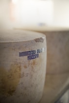 A large wheel of cheese with a textured surface is prominently displayed. The side of the cheese features a dark blue stamp with dates printed on it, indicating a likely aging process. The background is softly blurred, creating a focus on the cheese.