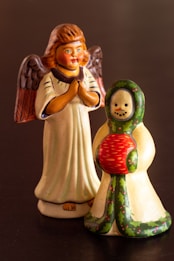 Two wooden figurines are displayed on a dark surface. On the left, there is an angelic figurine with carved wings, a white and brown robe, and a serene expression with hands clasped in front. On the right, a snowman figure is adorned with a green and red holly-patterned scarf or wrap, holding a red object, possibly a book. The figures are painted with vibrant colors and exhibit a handcrafted feel.