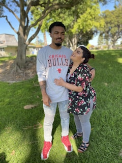 A couple stands together on a grassy area with trees in the background. The man is wearing a gray Chicago Bulls shirt, light blue jeans, and red and white sneakers. The woman is gazing up at him, wearing a floral top, gray leggings, and sandals. Their expressions suggest a mix of affection and humor.