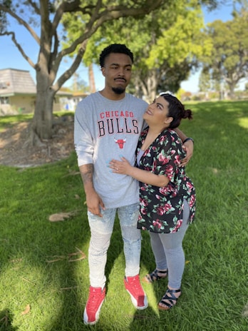 A couple stands together on a grassy area with trees in the background. The man is wearing a gray Chicago Bulls shirt, light blue jeans, and red and white sneakers. The woman is gazing up at him, wearing a floral top, gray leggings, and sandals. Their expressions suggest a mix of affection and humor.