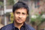 A young man with short dark hair is wearing a dark-colored jacket. He appears to be outdoors with a blurred background that includes some greenery and a building.