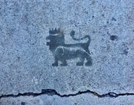 A black stencil of a lion wearing a crown is depicted on a textured concrete surface. The lion is standing in profile with its tail curled upwards.