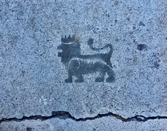 A black stencil of a lion wearing a crown is depicted on a textured concrete surface. The lion is standing in profile with its tail curled upwards.