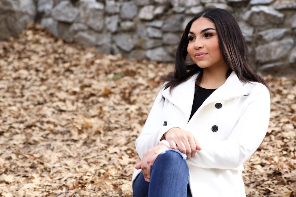 woman in white coat and blue denim jeans sitting on brown dried leaves during daytime