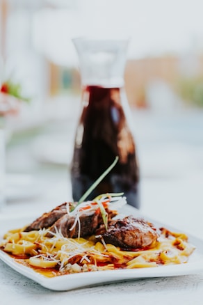 A dish featuring grilled meat placed on a bed of pasta, garnished with thinly sliced green onions and shredded cheese. The meal is presented on a white rectangular plate, with a blurred background showing a carafe filled with a dark liquid.