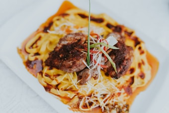A dish served on a square white plate, featuring two pieces of grilled meat placed on top of a bed of fettuccine pasta. The dish is garnished with sliced green onions, thin slivers of red chili, and shredded cheese. There is a rich sauce drizzled over the pasta.