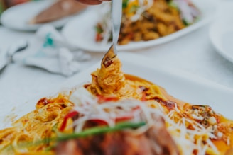 A fork is lifting a portion of a creamy, colorful dish, rich in spices and garnished with herbs and sliced vegetables. The surrounding environment suggests a dining setting with similar dishes blurred in the background.