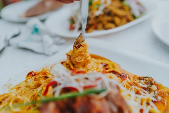 A fork is lifting a portion of a creamy, colorful dish, rich in spices and garnished with herbs and sliced vegetables. The surrounding environment suggests a dining setting with similar dishes blurred in the background.