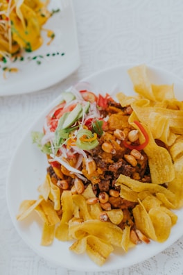 A plate of food featuring crispy plantain chips, garnished with sliced vegetables such as cucumber, tomato, and red onion. Roasted peanuts and what appears to be a meat-based dish are also present, adding texture and flavor. The food is arranged on a white plate with a decorative pattern on the tablecloth underneath.