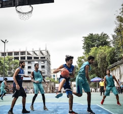 Players engaged in a competitive game of basketball on an outdoor court, surrounded by trees and a tall, partially constructed building in the background.