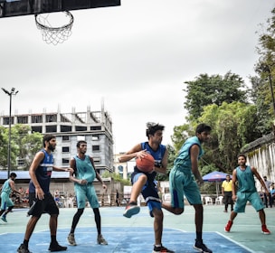 Players engaged in a competitive game of basketball on an outdoor court, surrounded by trees and a tall, partially constructed building in the background.