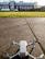 A small white drone is positioned on a textured concrete surface with a large building in the background. The building features multiple floors with large windows and red brick accents. In the foreground, there is a grassy lawn that separates the drone from the building. The sky is partly cloudy, and the scene is well-lit by sunlight.