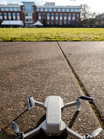 A small white drone is positioned on a textured concrete surface with a large building in the background. The building features multiple floors with large windows and red brick accents. In the foreground, there is a grassy lawn that separates the drone from the building. The sky is partly cloudy, and the scene is well-lit by sunlight.
