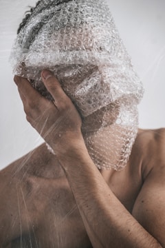 A person with their face covered in bubble wrap, one hand touching the side of their face, creating a surreal and thought-provoking visual composition.