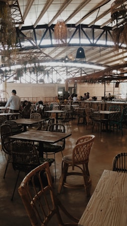 A spacious, rustic cafe or restaurant with numerous wooden tables and wicker chairs arranged across the floor. The ceiling is high with visible beams, adorned with hanging plants and light fixtures. Natural light filters through large windows, illuminating the earthy tones of the interior. Several patrons are seated, engaged in conversation.