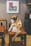A cat with a mix of brown, black, and white fur is sitting on a wooden stool, grooming itself. The backdrop includes a sign with a colorful logo and some furniture, creating a cozy setting.