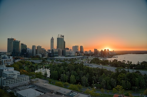 The image features a city skyline at sunset, with skyscrapers silhouetted against a vibrant orange sky. The sun is setting on the horizon, casting a warm glow over the buildings. Below, there is a mix of urban and natural landscape, including green trees and a body of water.