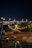 A nighttime scene featuring a warmly lit outdoor patio with string lights hanging above. In the foreground, there is a small pool with a jacuzzi, bordered by stone seating areas. Beyond the patio, a cityscape brightly lights the horizon, blending with the night sky.