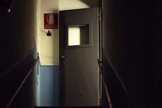 A dimly lit corridor features a dark gray door with a small square window. The walls are primarily blue with a white upper section. A red rectangular fire extinguisher sign with the word 'ESTINTORE' is visible on the wall.