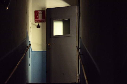 A dimly lit corridor features a dark gray door with a small square window. The walls are primarily blue with a white upper section. A red rectangular fire extinguisher sign with the word 'ESTINTORE' is visible on the wall.