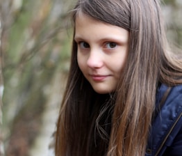 A young girl with long brown hair and a subtle smile glances sideways. She is wearing a dark blue jacket, and the background features blurred natural scenery, likely a forest or park setting.