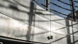 A modern architectural structure with a curved metallic wall and large windows. The shadows of window frames create patterns on the wall. A small speaker or light fixture is mounted on the lower section of the wall.