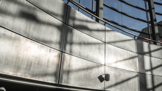 A modern architectural structure with a curved metallic wall and large windows. The shadows of window frames create patterns on the wall. A small speaker or light fixture is mounted on the lower section of the wall.