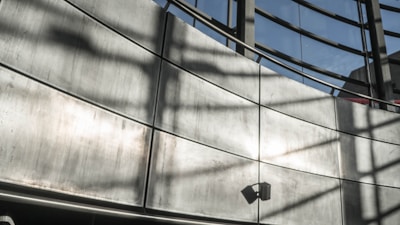 A modern architectural structure with a curved metallic wall and large windows. The shadows of window frames create patterns on the wall. A small speaker or light fixture is mounted on the lower section of the wall.