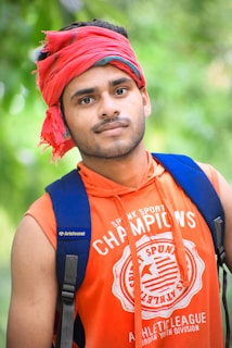 A young person wearing an orange sleeveless shirt with a logo and text, carrying a blue backpack. A red headscarf is wrapped around the head. The background is blurred with green foliage, suggesting an outdoor setting.