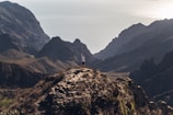 A solitary person stands triumphantly on a rugged mountain peak, arms raised against a backdrop of dramatic, shadowed cliffs and the distant, shimmering sea. The landscape is vast and majestic, capturing a sense of freedom and achievement.