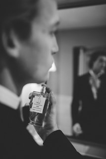 A black and white image of a person seen in profile holding a bottle of perfume near their face, with the reflection of another person visible in the background mirror. The focus is on the hand and the perfume bottle labeled 'Picci 540', with the rest of the image slightly blurred.