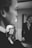 A black and white image of a person seen in profile holding a bottle of perfume near their face, with the reflection of another person visible in the background mirror. The focus is on the hand and the perfume bottle labeled 'Picci 540', with the rest of the image slightly blurred.