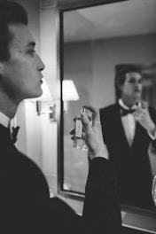 A man in formal attire, likely a tuxedo, stands in front of a mirror. He is holding a bottle of perfume close to his face, as if preparing to apply it. The setting appears to be a well-lit bathroom with a visible wall lamp.
